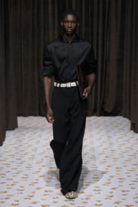 Male model walking a runway in a black satin shirt, black trousers, a white belt, and a brown clutch under dark drapes.