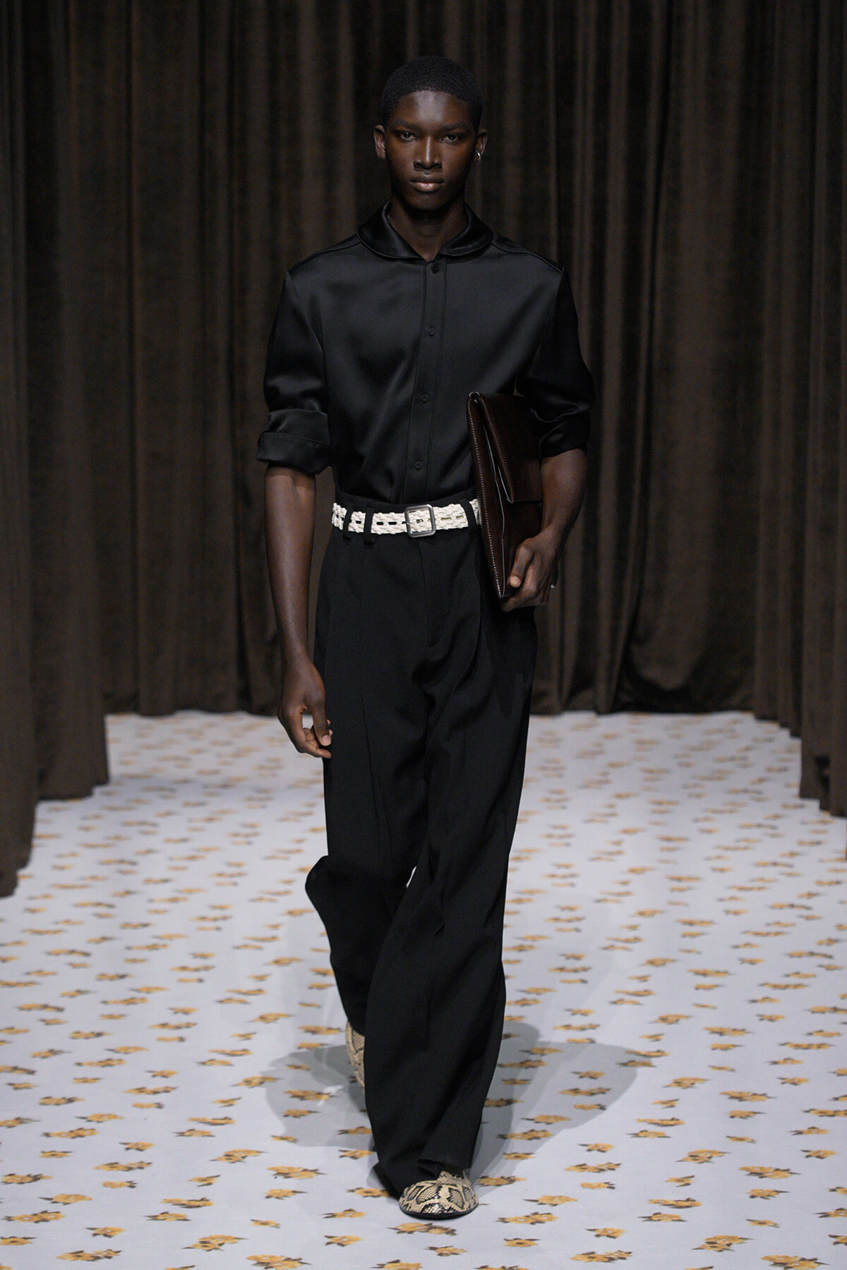 Male model walking a runway in a black satin shirt, black trousers, a white belt, and a brown clutch under dark drapes.