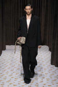 Male model walking a fashion runway in a black oversized suit with a pale pink shirt, carrying a snakeskin-patterned bag.