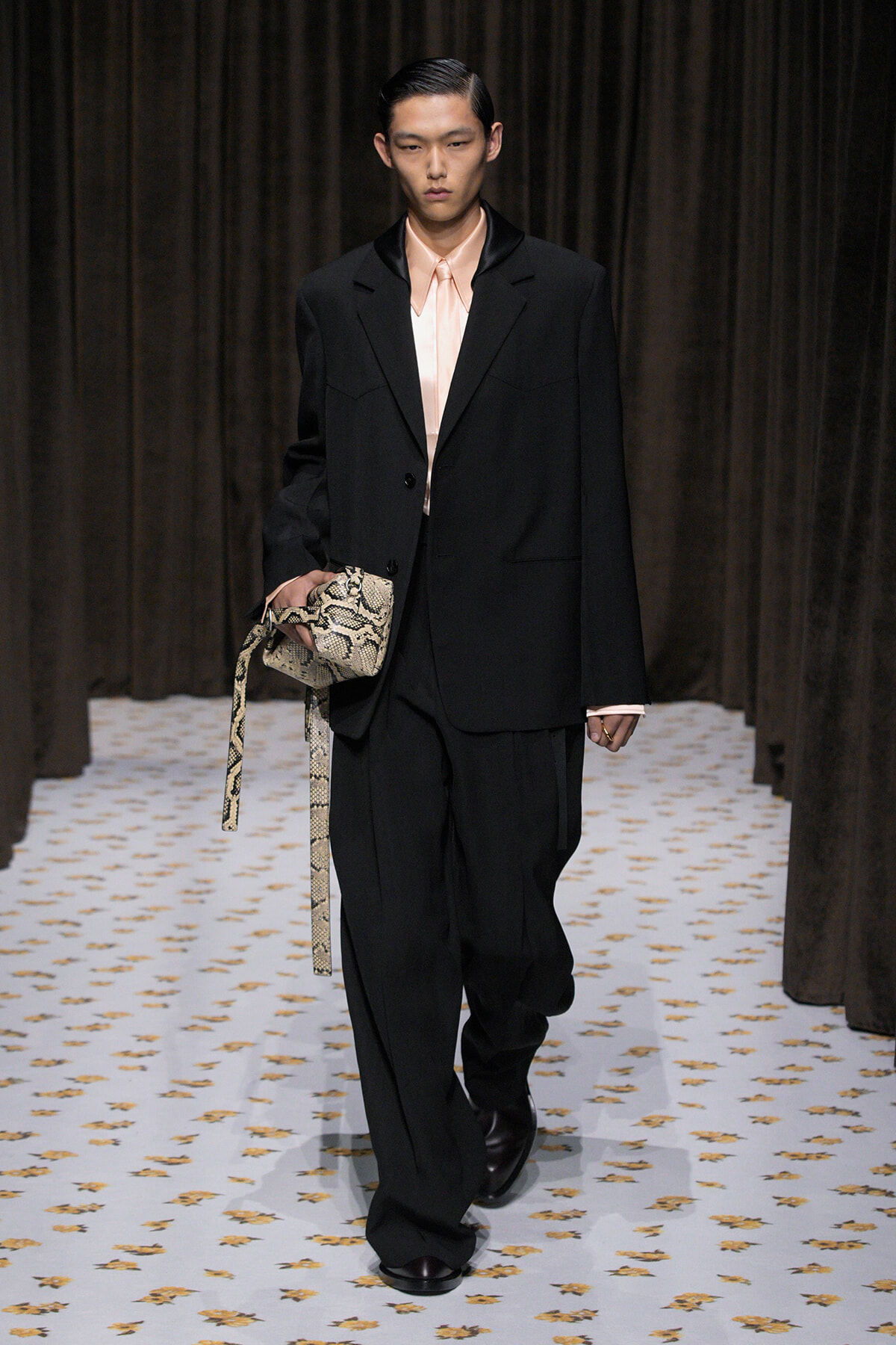 Male model walking a fashion runway in a black oversized suit with a pale pink shirt, carrying a snakeskin-patterned bag.