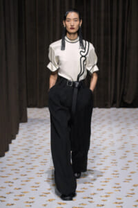 Model walks the runway in a white top with black piping and wide black trousers belted at the waist.