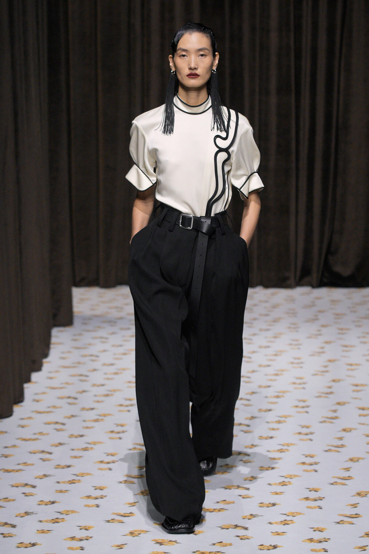 Model walks the runway in a white top with black piping and wide black trousers belted at the waist.