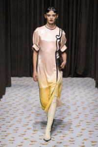 Model walks the runway in a satin gradient dress from pink to yellow with black piping, carrying a clutch.