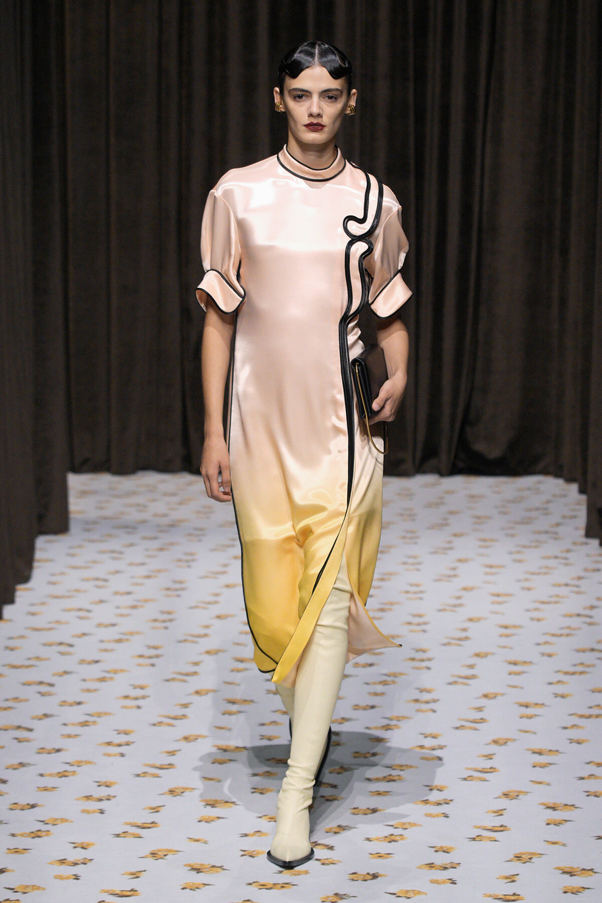 Model walks the runway in a satin gradient dress from pink to yellow with black piping, carrying a clutch.