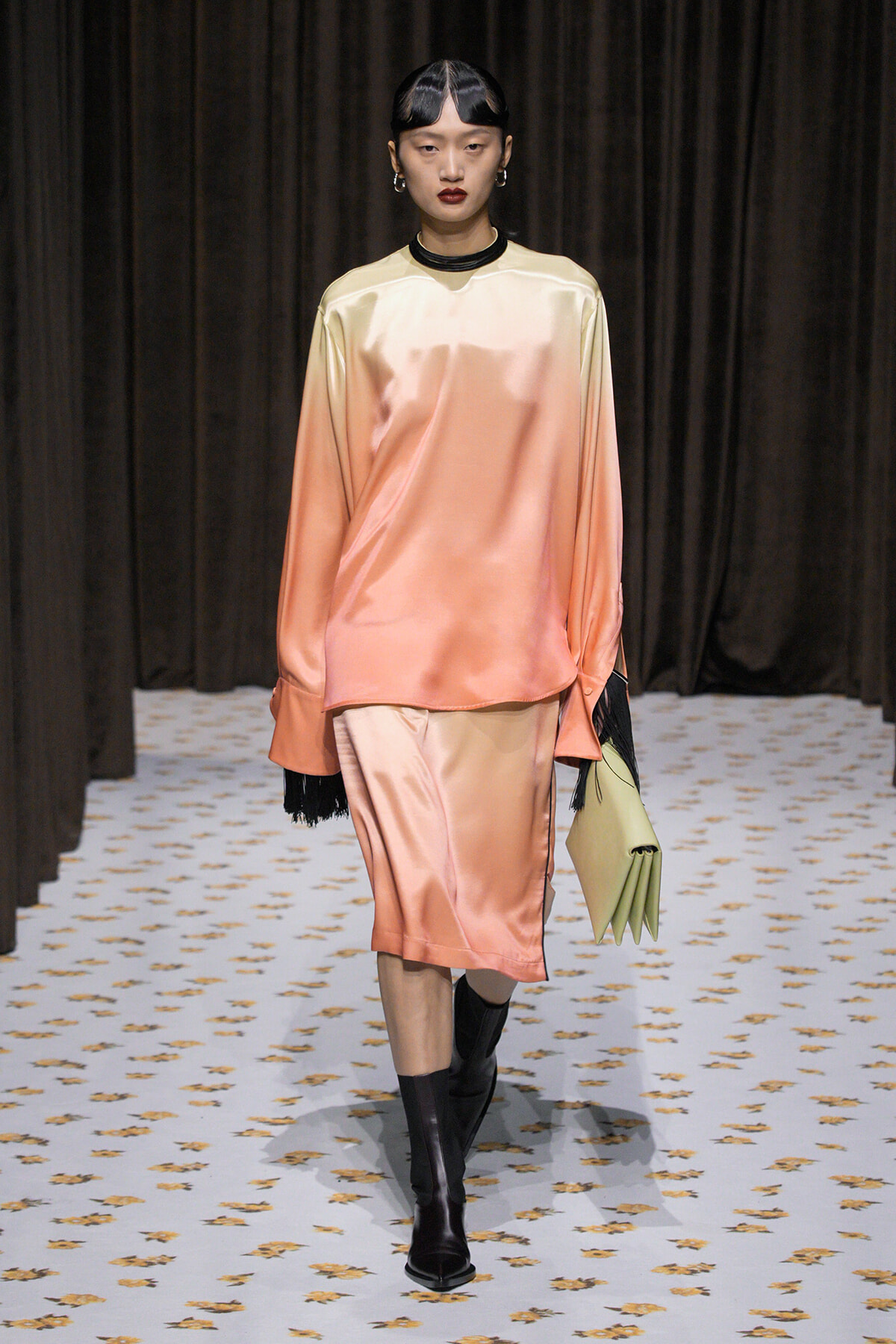 Model wearing a peach-pink satin gradient tunic and matching knee-length skirt, black knee-high boots, and a pale green clutch, walking a runway with dark curtains in the background.