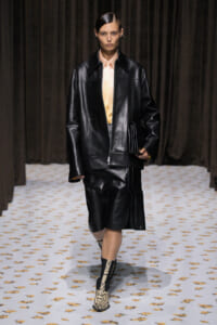 Model walking runway in oversized black leather jacket, matching skirt, pale peach blouse, and snakeskin boots, holding a black clutch.