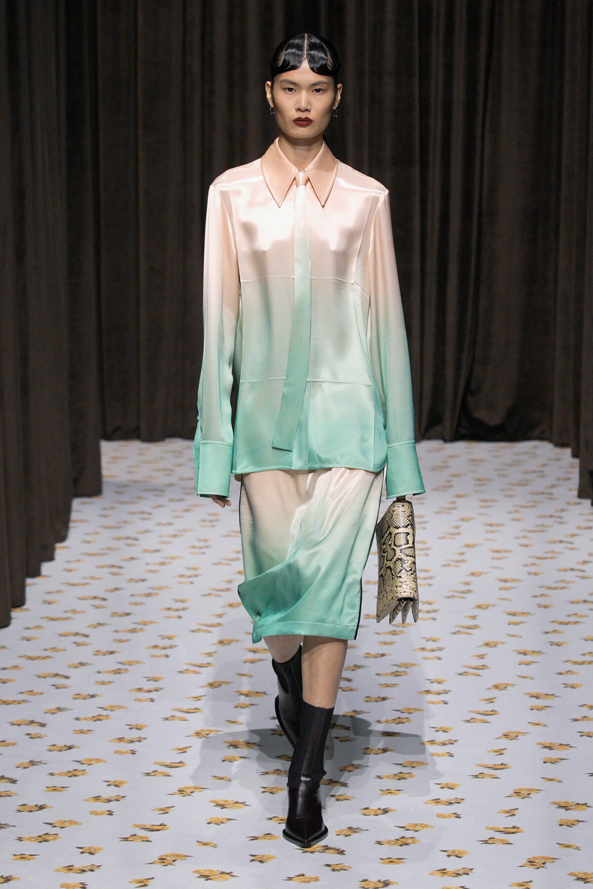 Model wearing a peach-to-mint gradient satin blouse and matching skirt on the runway, holding a snakeskin clutch.