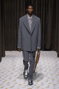 Male model walking a runway in a structured gray suit with oversized lapels, pale shirt, and a brown leather briefcase.
