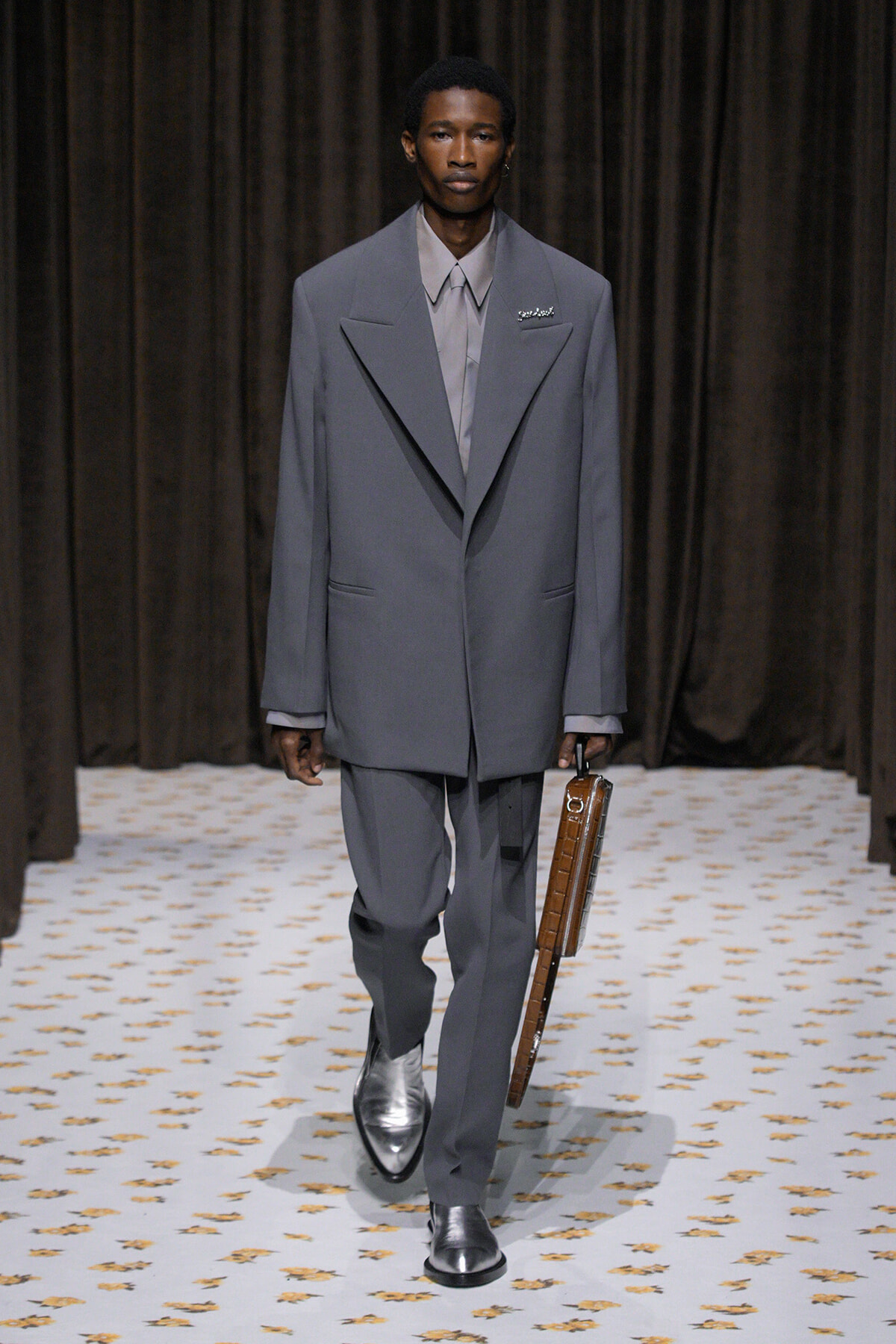 Male model walking a runway in a structured gray suit with oversized lapels, pale shirt, and a brown leather briefcase.