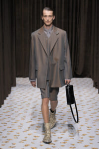 Male model on a runway wearing an oversized gray suit with matching shorts, snakeskin boots, and a black briefcase.