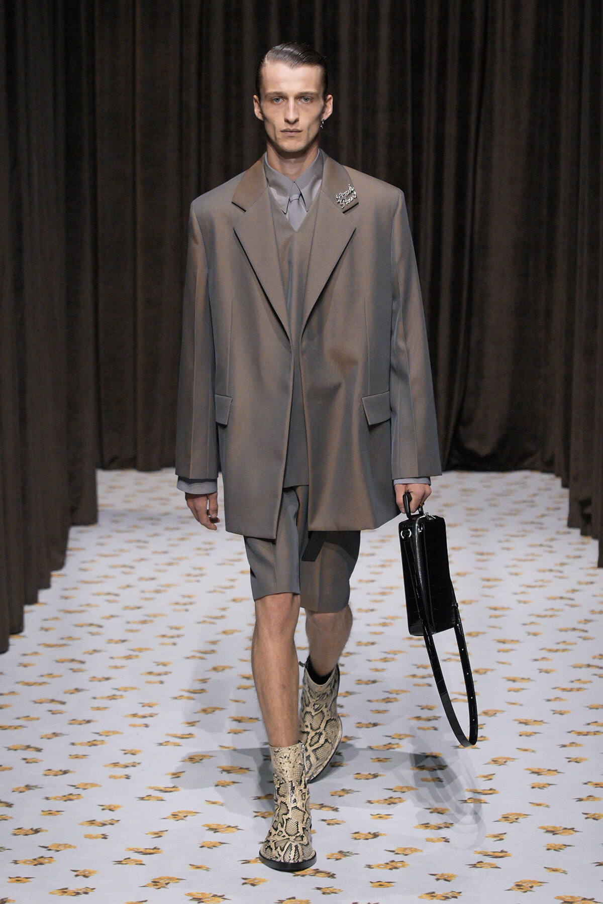 Male model on a runway wearing an oversized gray suit with matching shorts, snakeskin boots, and a black briefcase.