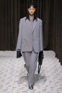 Model walking the runway in an oversized light gray suit with a matching tie, holding a black handbag against a dark backdrop.