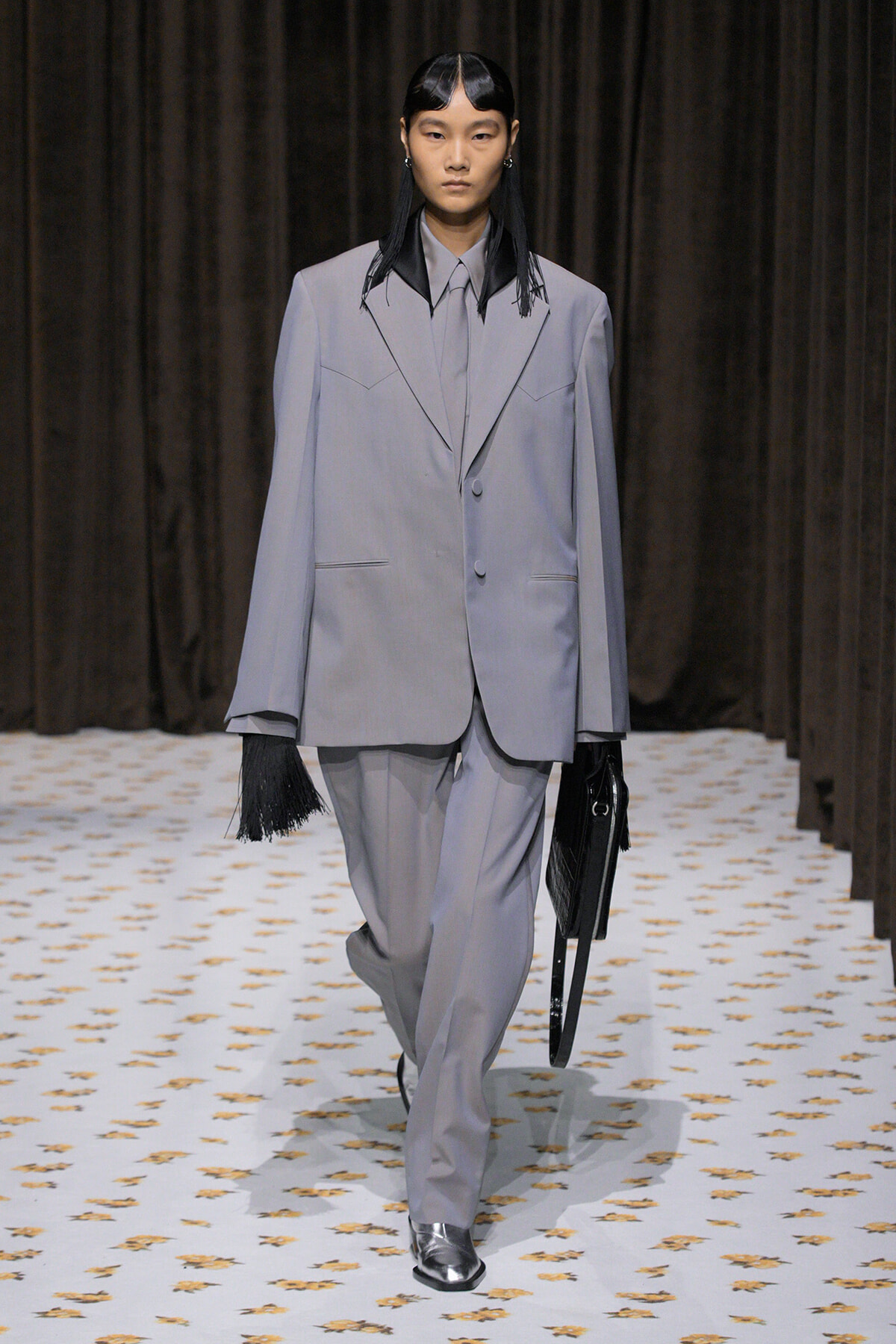 Model walking the runway in an oversized light gray suit with a matching tie, holding a black handbag against a dark backdrop.