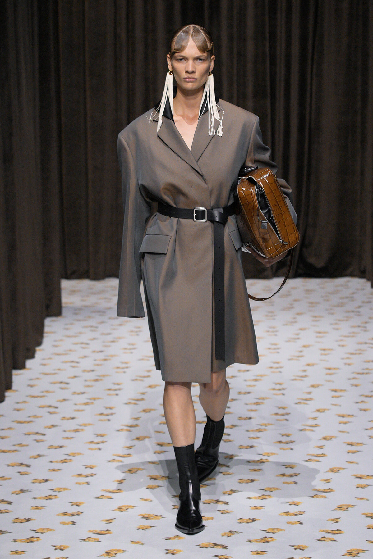 Model on a runway wearing a gray belted wrap coat, black boots, and bold white fringe earrings, carrying a brown croc-embossed handbag.