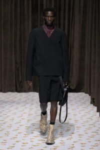 Male model on a runway wearing an oversized black V-neck sweater over a burgundy collared shirt, black shorts, snakeskin boots, and a black bag.