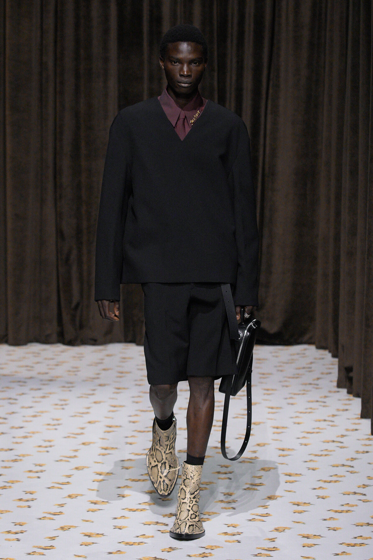 Male model on a runway wearing an oversized black V-neck sweater over a burgundy collared shirt, black shorts, snakeskin boots, and a black bag.