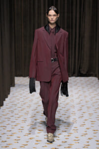 Model walking a burgundy suit with a black satin lapel, burgundy shirt, and belt on a patterned runway; black fringe gloves add drama.