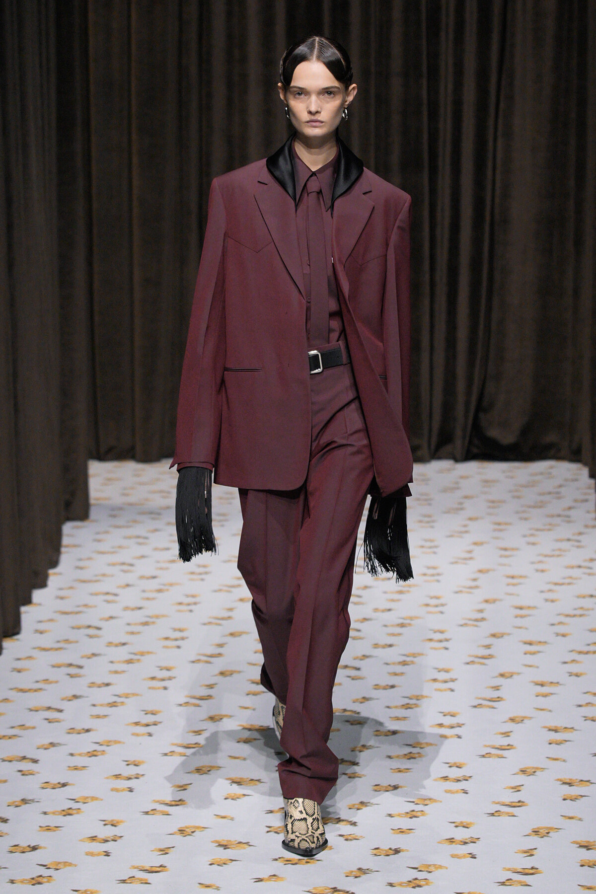 Model walking a burgundy suit with a black satin lapel, burgundy shirt, and belt on a patterned runway; black fringe gloves add drama.