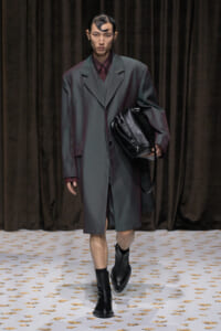 Male model on a runway wearing an oversized charcoal-gray coat over a burgundy suit, carrying a black leather bag and wearing black boots.