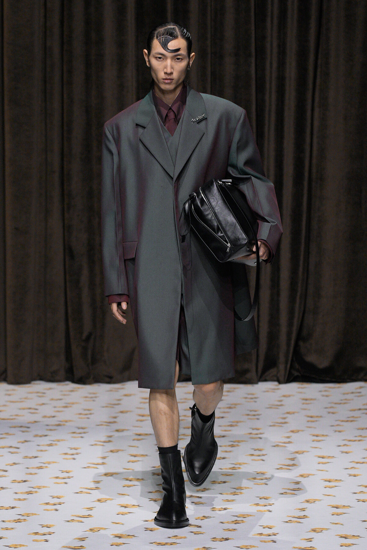 Male model on a runway wearing an oversized charcoal-gray coat over a burgundy suit, carrying a black leather bag and wearing black boots.