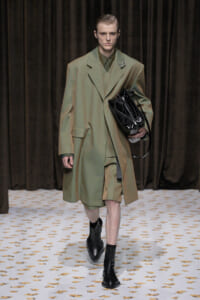 Male model walking a fashion runway in an oversized olive-green coat, matching suit, and black boots, carrying a black patent handbag.