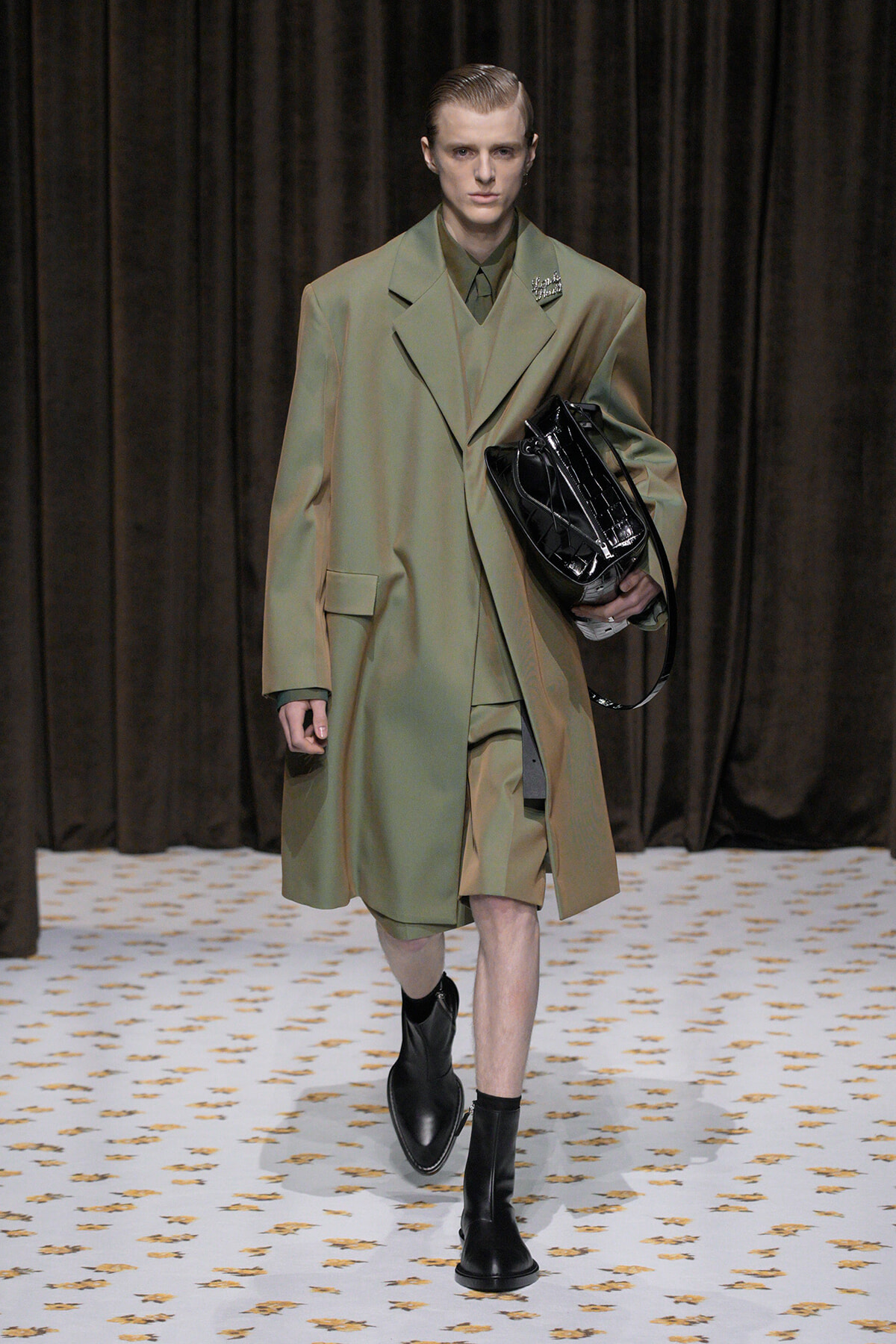 Male model walking a fashion runway in an oversized olive-green coat, matching suit, and black boots, carrying a black patent handbag.