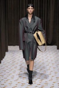 Model walks a runway in an oversized gray belted coat, carrying a beige structured bag and wearing black ankle boots beside dark curtains.