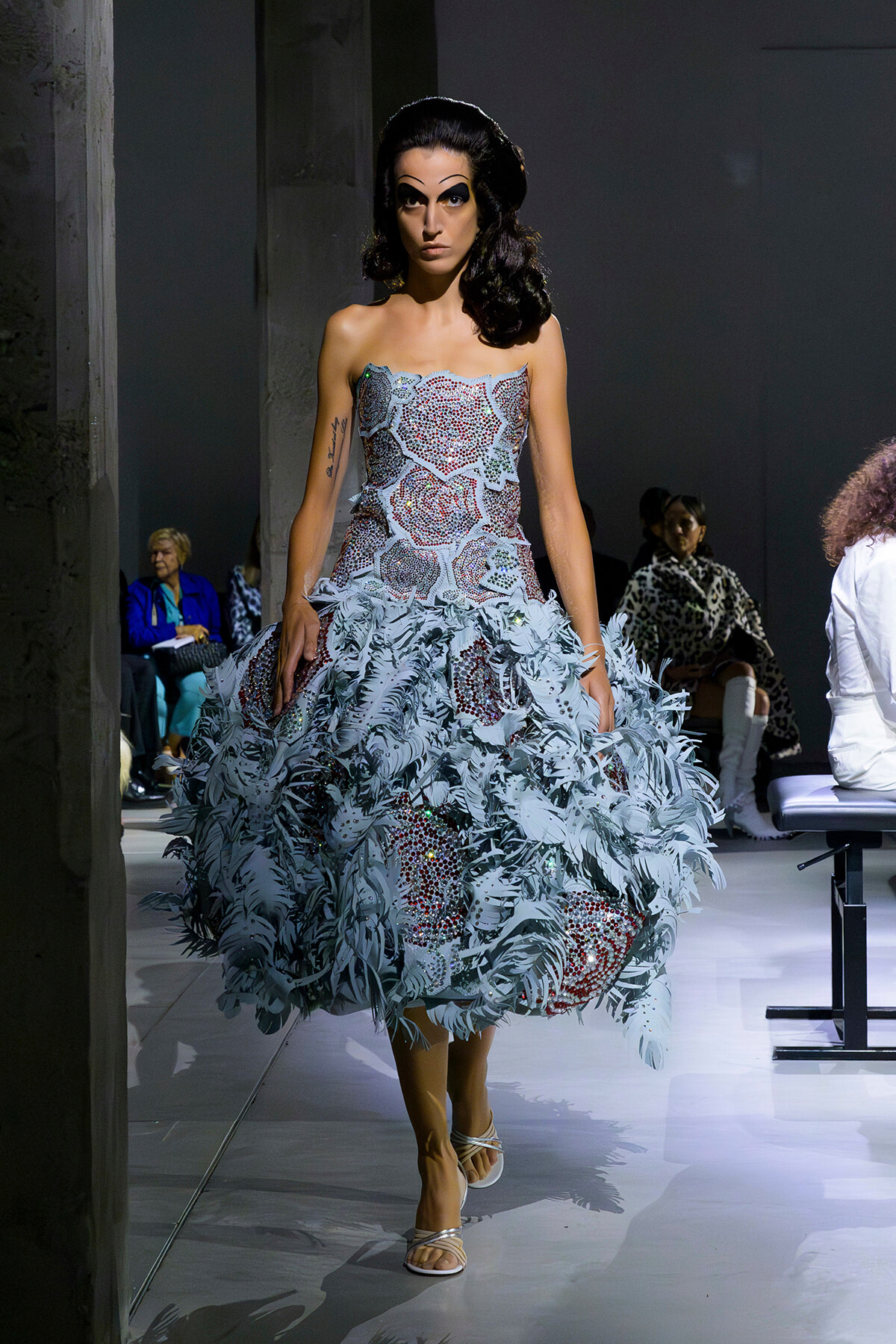 Model walking the runway in a strapless, beaded gown with a voluminous pale blue feathered skirt and white sandals.
