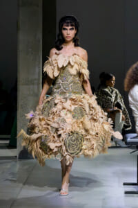 Runway model in a beige feathered off-shoulder couture gown with intricate beadwork walking a fashion show.