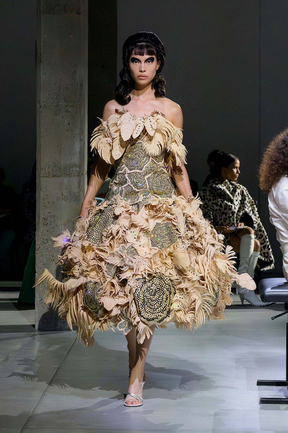 Runway model in a beige feathered off-shoulder couture gown with intricate beadwork walking a fashion show.