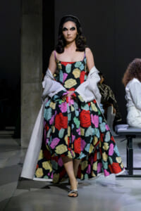 Model walking a fashion runway in a bold, multicolored rose-print gown with a white cape draped over the shoulders.