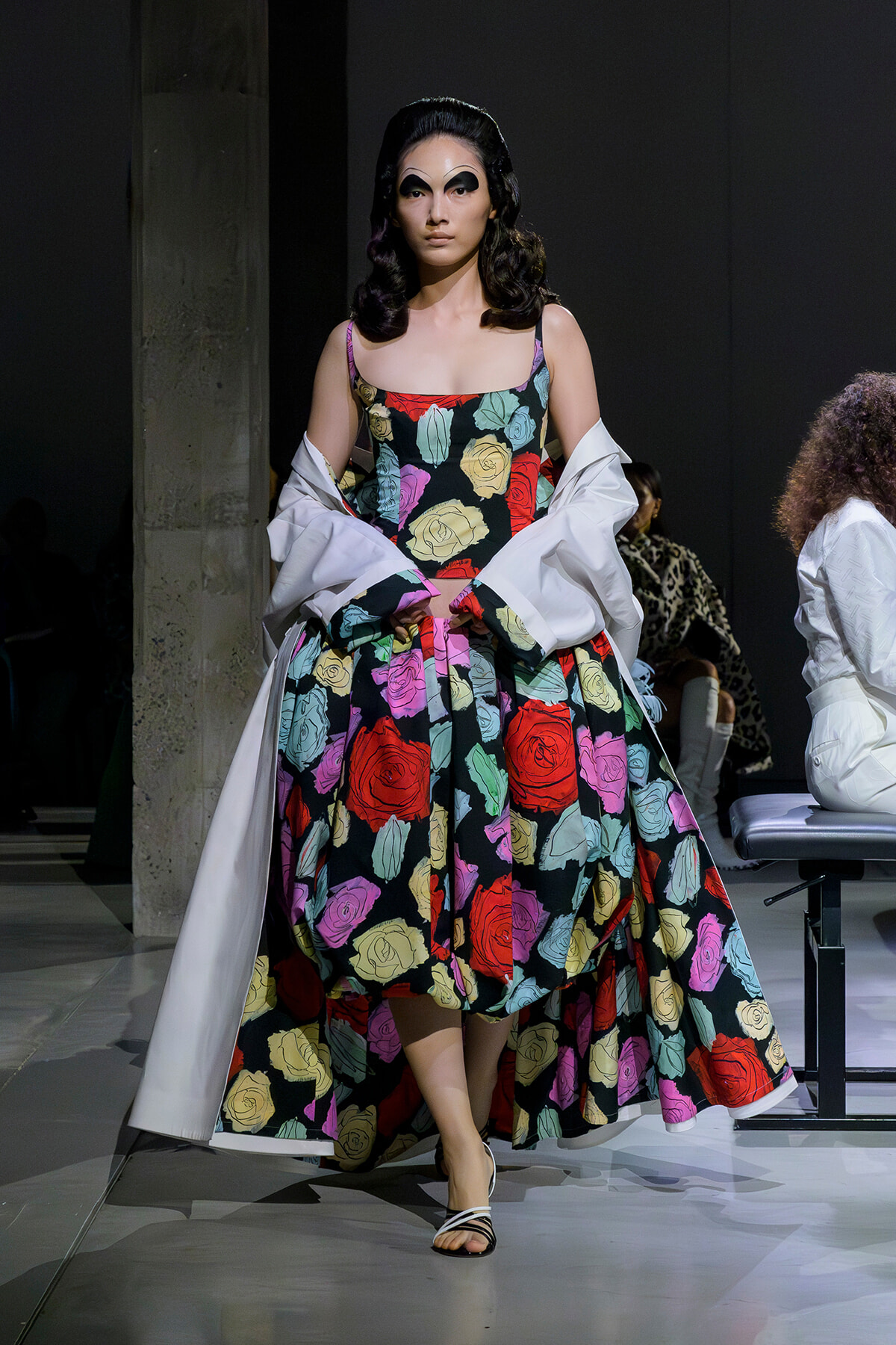 Model walking a fashion runway in a bold, multicolored rose-print gown with a white cape draped over the shoulders.