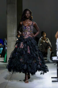 Model walking the runway in a voluminous black feathered gown with a beaded floral bodice; audience in background at a fashion show