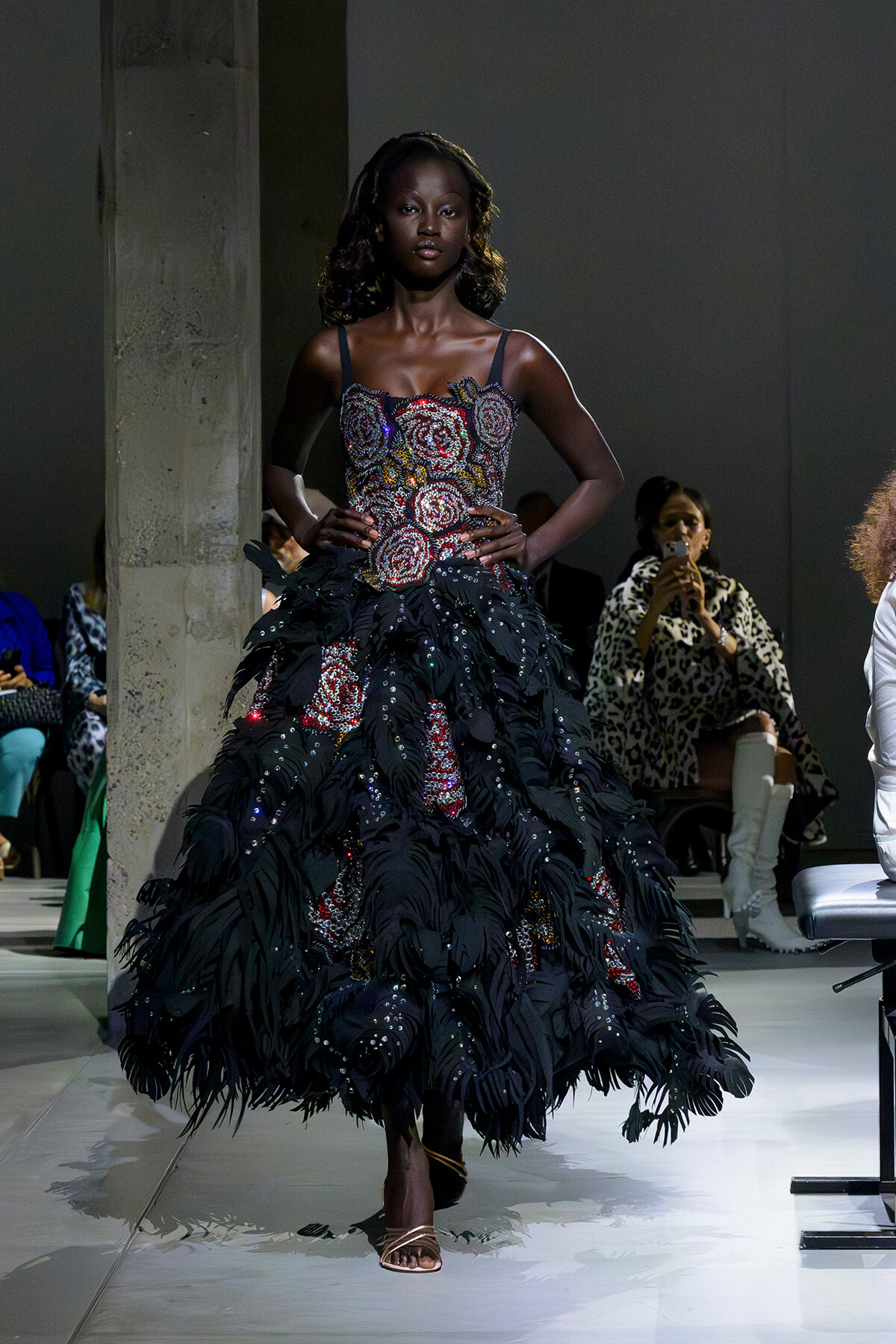 Model walking the runway in a voluminous black feathered gown with a beaded floral bodice; audience in background at a fashion show