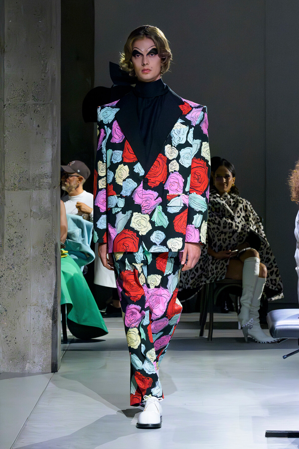 Model walks a runway in a bold floral suit with oversized shoulders and black turtleneck, dramatic face makeup.