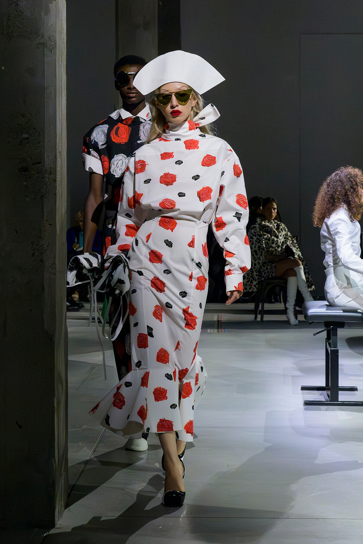 Model walking a fashion runway in a white rose-print dress with a large white sculptural hat and yellow sunglasses.