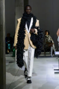 Male model walking a runway in a beige blazer, white pants, and an oversized black feathered scarf draped over the shoulders.