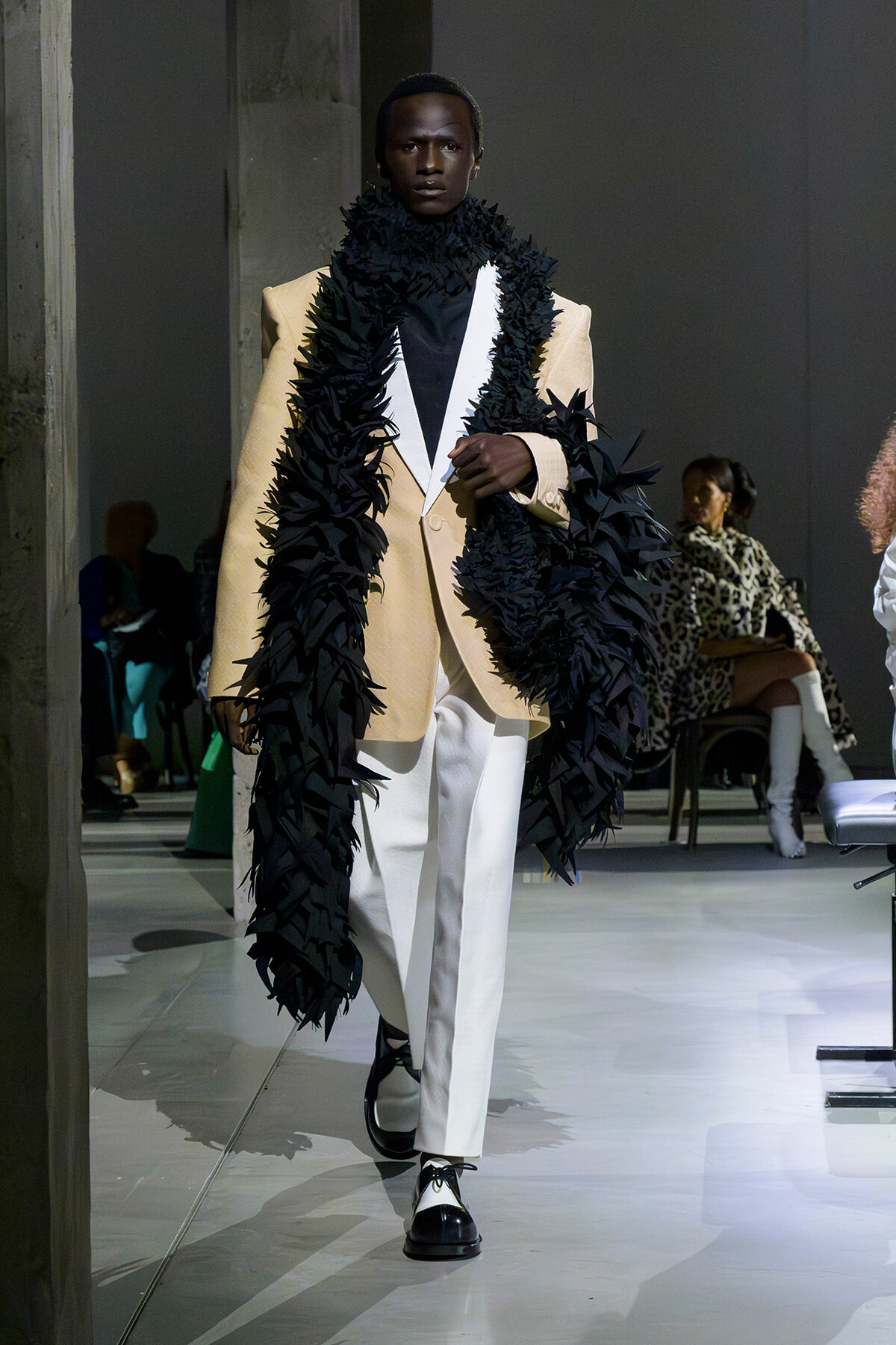 Male model walking a runway in a beige blazer, white pants, and an oversized black feathered scarf draped over the shoulders.