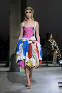 Model walking a runway in a bold, multicolor dress with a pink top and large abstract floral skirt, blond hair and dramatic makeup.