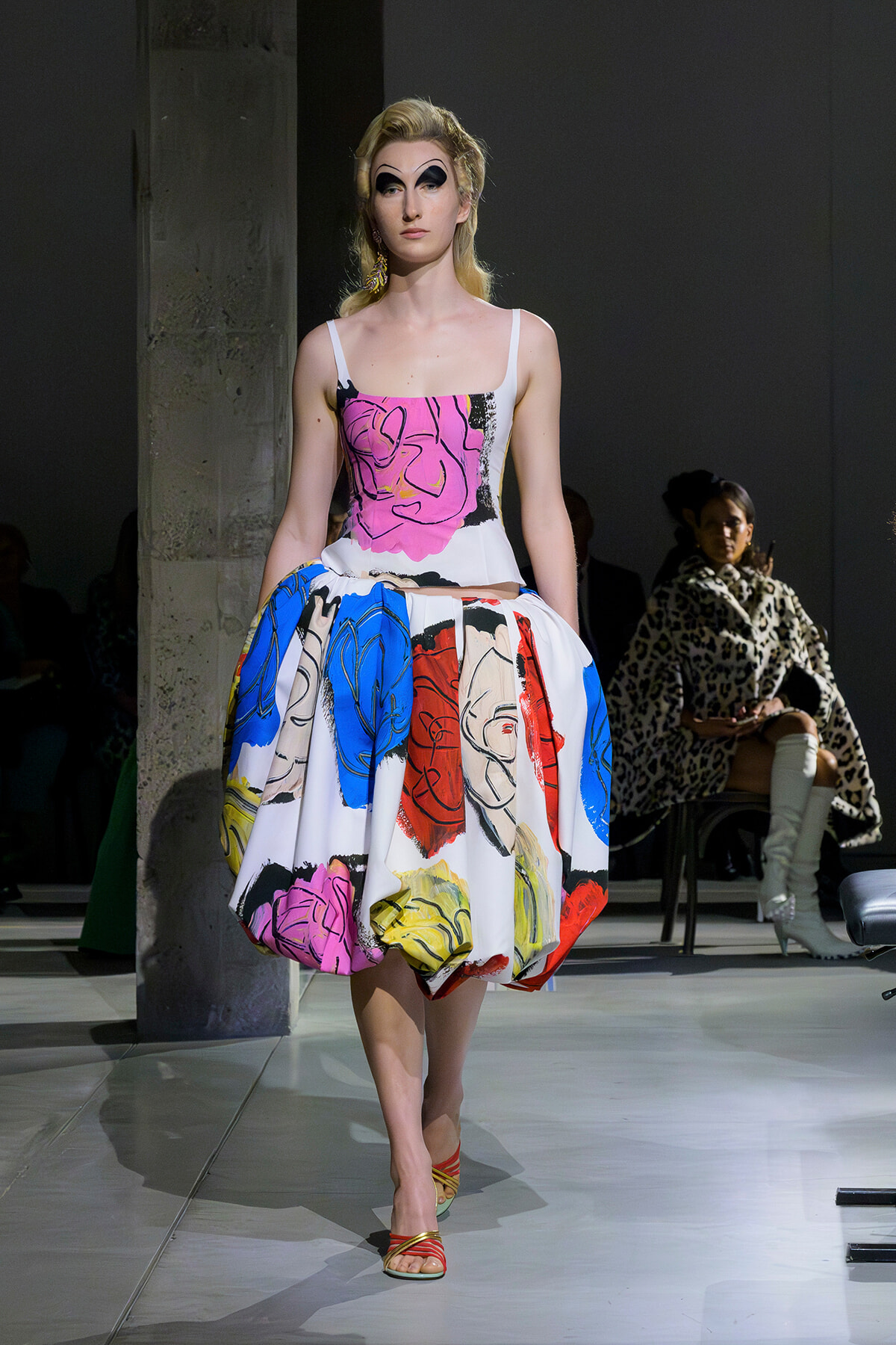 Model walking a runway in a bold, multicolor dress with a pink top and large abstract floral skirt, blond hair and dramatic makeup.