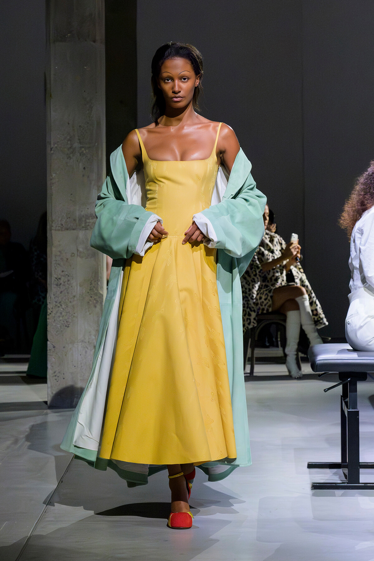 Model walking the runway in a pale yellow dress with a mint green outer coat, red shoes, and bold styling under studio lights.