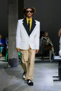 Male model on a runway wearing a white blazer with black lapels, a yellow shirt and tie, beige wide-leg pants, black-and-white shoes, and oversized sunglasses.