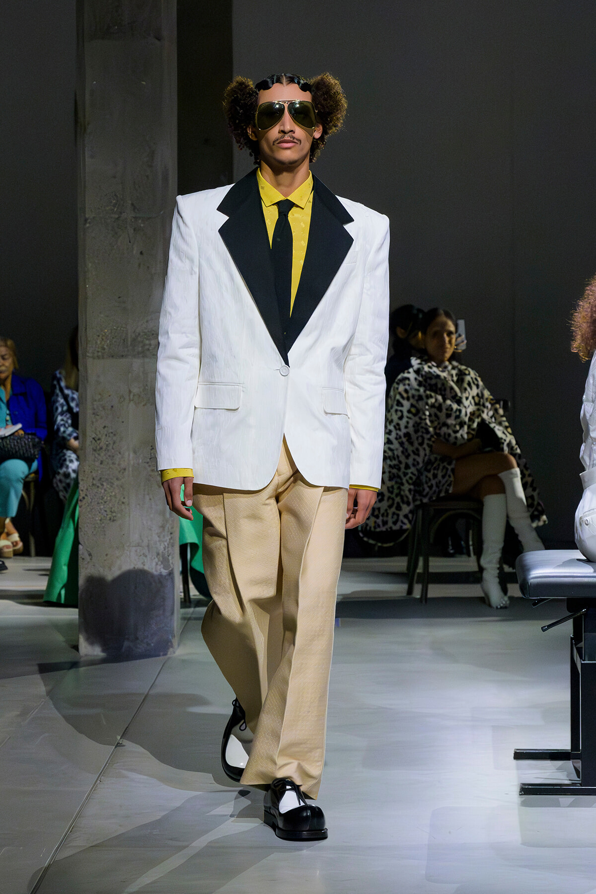 Male model on a runway wearing a white blazer with black lapels, a yellow shirt and tie, beige wide-leg pants, black-and-white shoes, and oversized sunglasses.