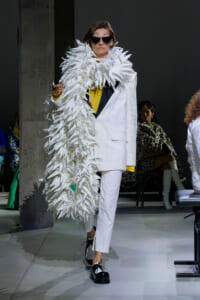 Model walks the runway in a white tuxedo with a dramatic white feather cape, yellow top peeking through, and oversized sunglasses.