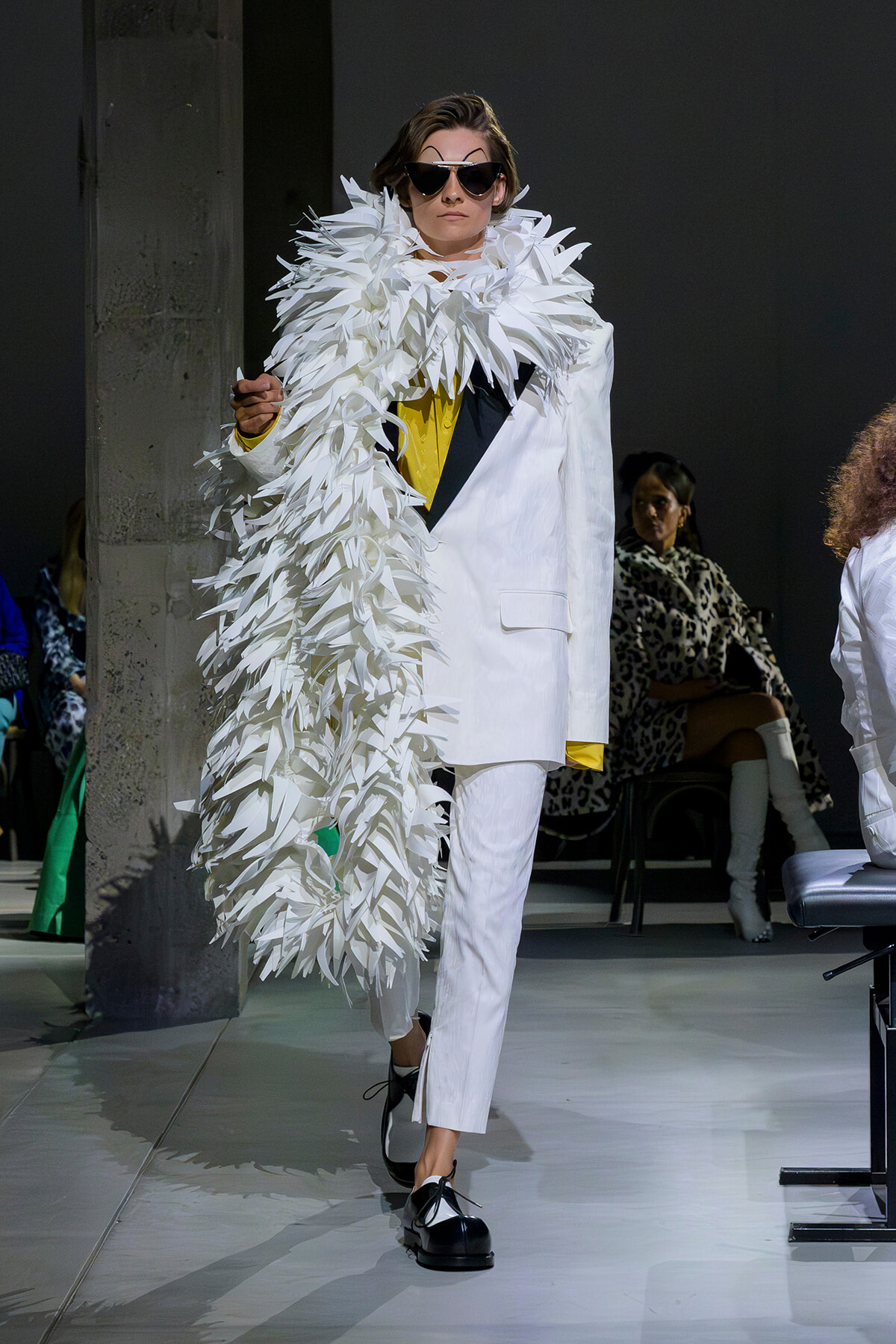 Model walks the runway in a white tuxedo with a dramatic white feather cape, yellow top peeking through, and oversized sunglasses.