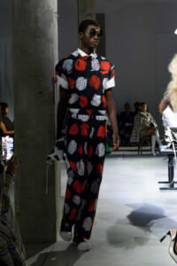 Model on a runway wearing a black floral jumpsuit with red and white roses, white collar, and oversized sleeves, paired with sunglasses and white shoes.