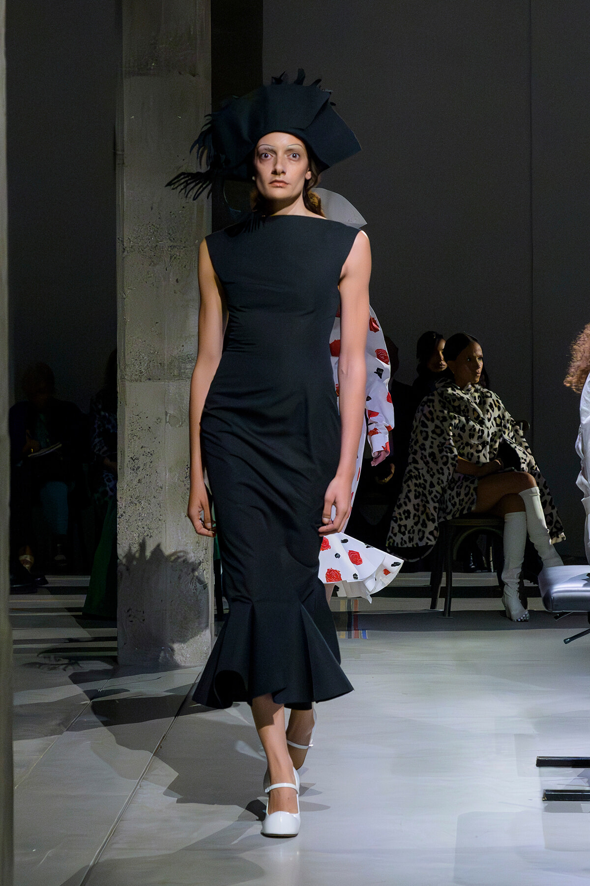 Model on the runway wearing a fitted black sleeveless mermaid dress with a large black hat and white heels.