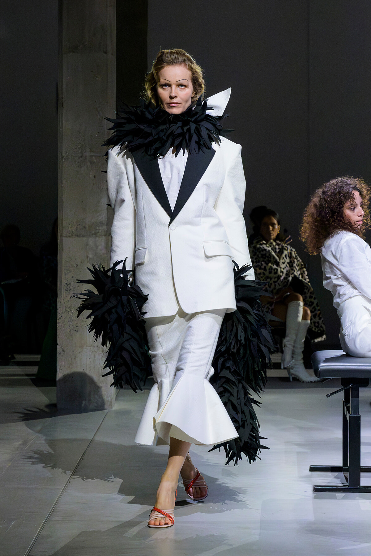 Model on a runway wearing a white suit with a dramatic black feather collar and cuffs, walking forward.