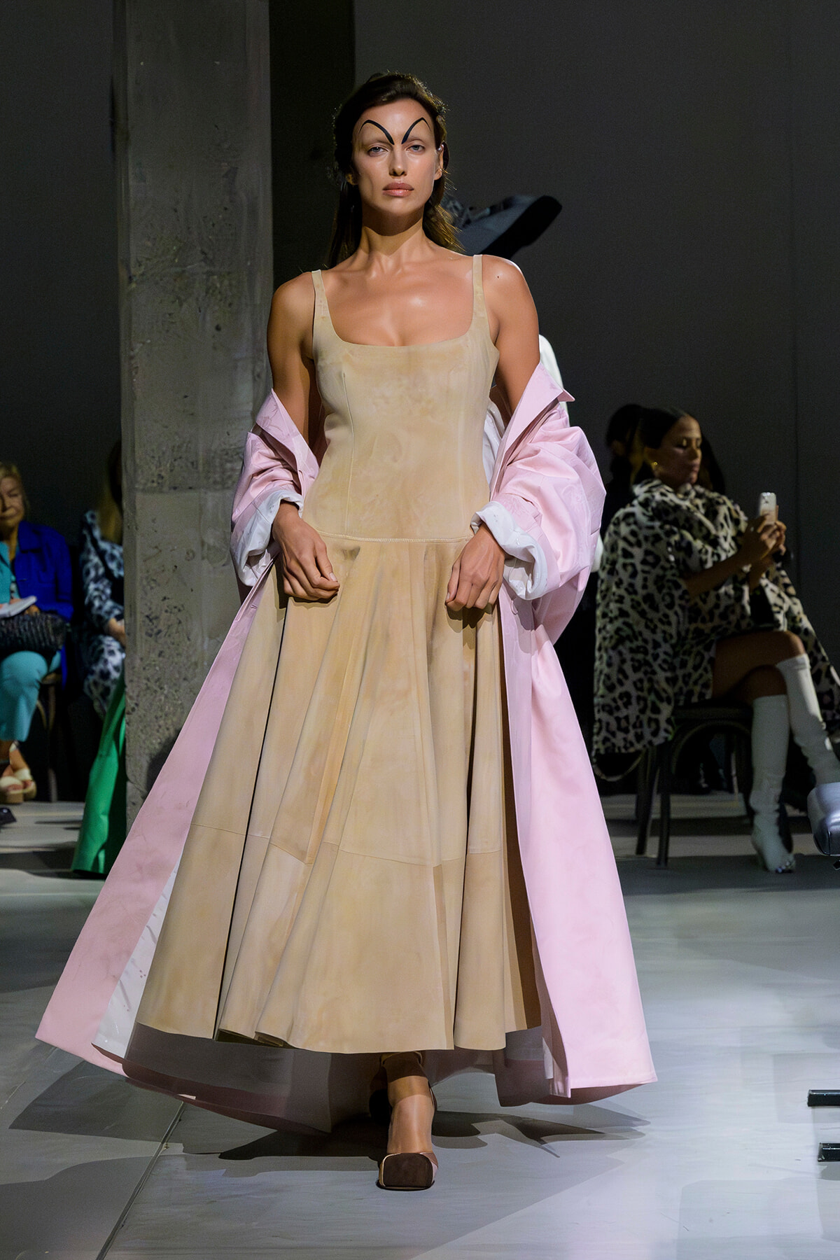 Model on a runway wearing a beige sleeveless gown with a pale pink coat draped over her shoulders.