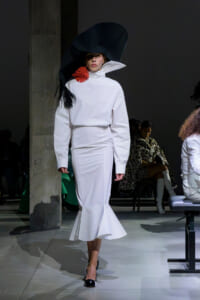 Model in a white high-neck outfit with an oversized black hat and a red feather accessory walking the runway.